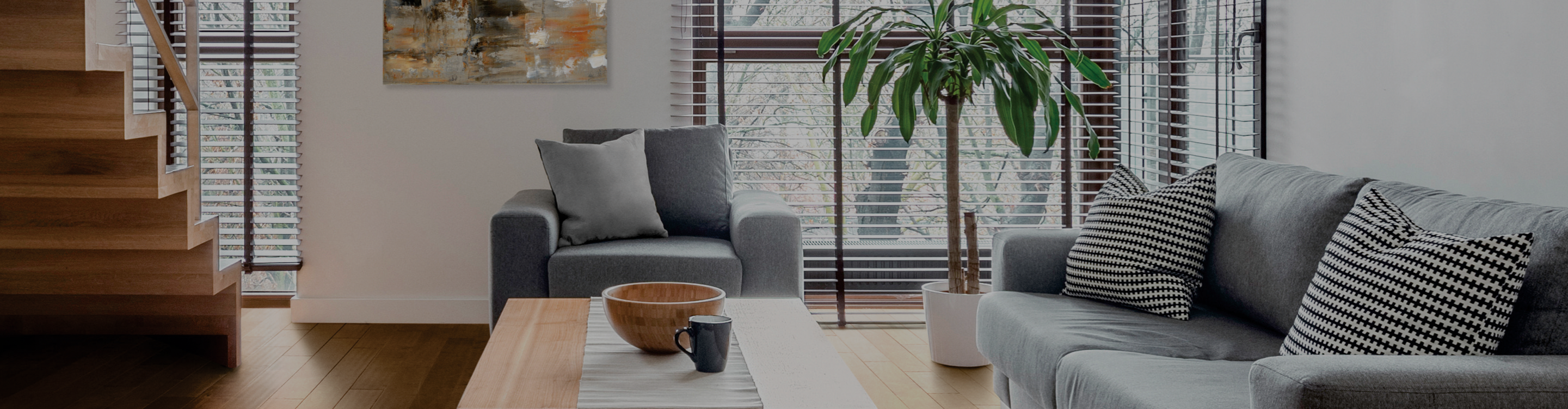 A modern living room with a grey sofa, armchair, wooden coffee table, wooden staircase, and a large potted plant near the window.